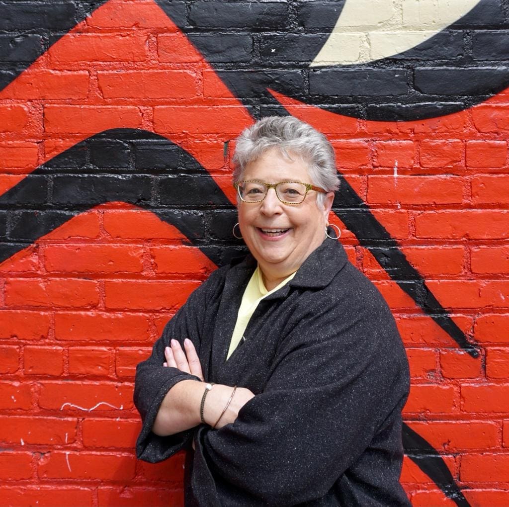A smiling Suzanne Goss stand in front of a bold black and red painted brick wall, with arms crossed.